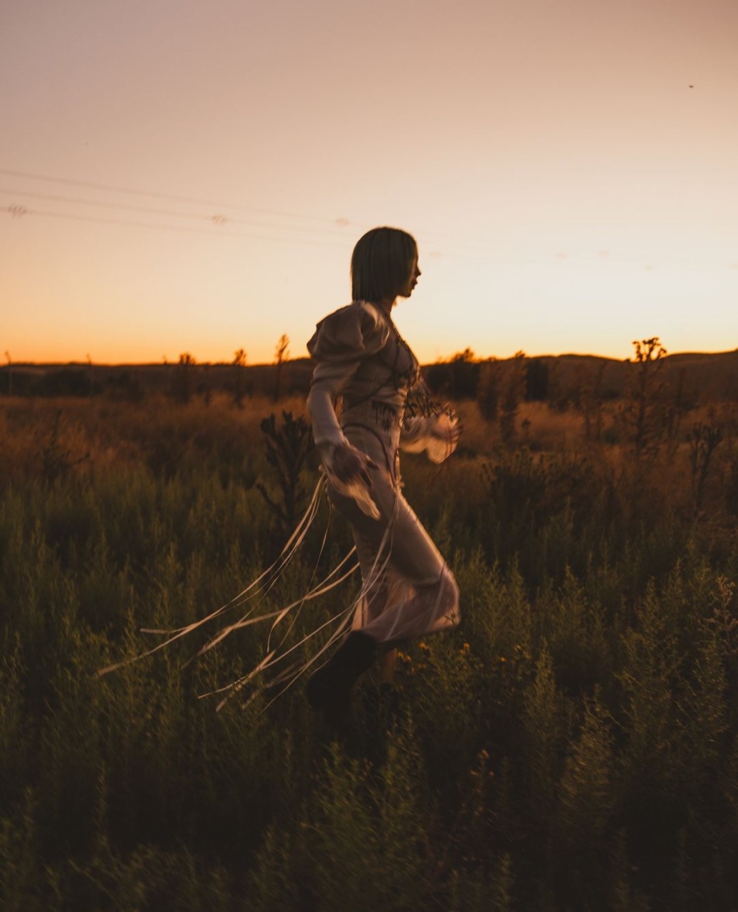 Adult female walking in a tall grassy field at sunset. Woman wears a vintage dress with flowing ribbons, walking away from the camera.