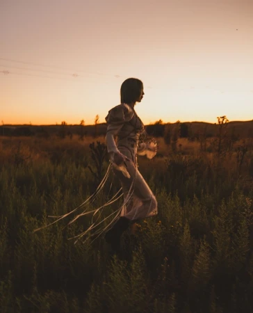 Adult female walking in a tall grassy field at sunset. Woman wears a vintage dress with flowing ribbons, walking away from the camera.