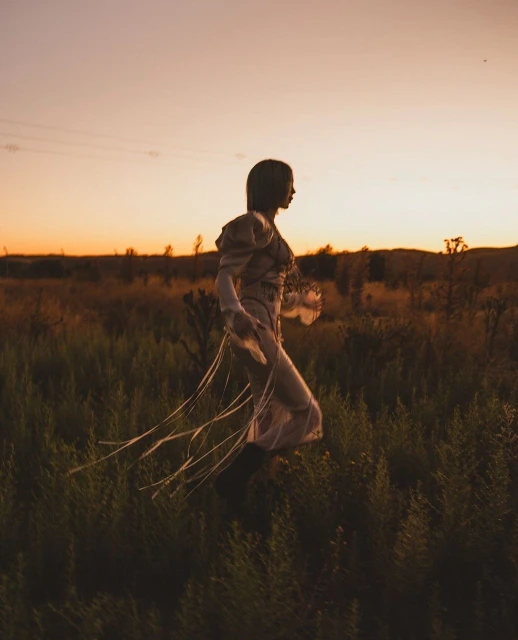 Woman in gown walks through field at sunset. She carries object and the golden hour lighting accentuates the tall grass.