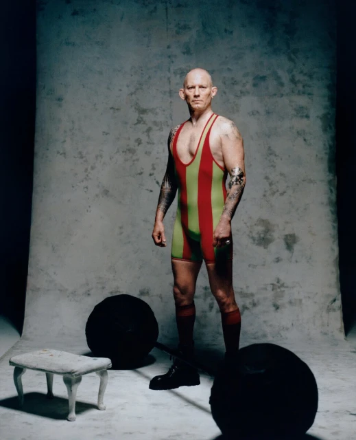 A muscular, bald adult male stands in a vintage striped wrestling singlet next to a barbell weight and an antique stool in a studio.