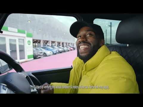 Smiling Black man in yellow jacket driving a car; parked cars and modern building visible through the window, reflecting natural light.