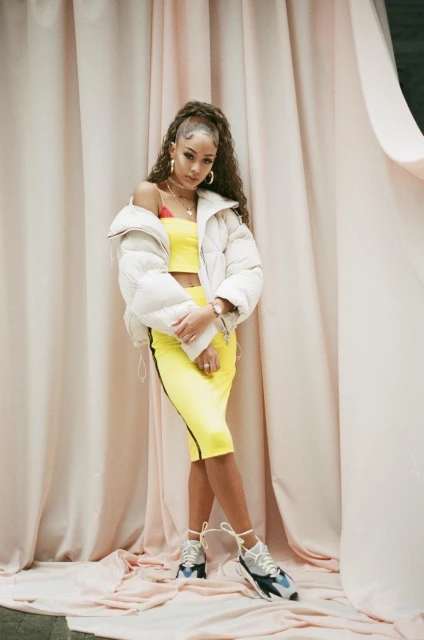 A stylish young woman with long, curly brown hair poses confidently in a yellow skirt and crop top with a white puffer jacket.