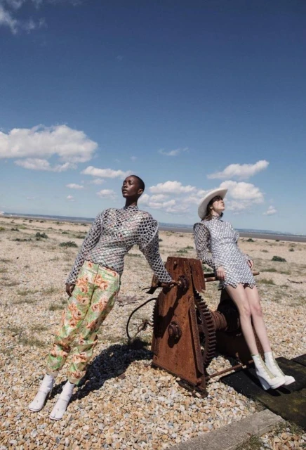 Two diverse women pose outdoors in artistic fashion, one in floral pants and a mirrored top, the other in a matching dress and hat.
