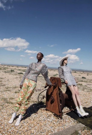 Two diverse women pose outdoors in artistic fashion, one in floral pants and a mirrored top, the other in a matching dress and hat.