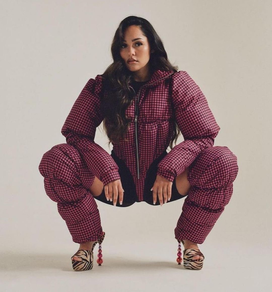 A woman squats in a studio wearing a puffy red and black houndstooth jacket with matching leg coverings and platform heels with beaded details.