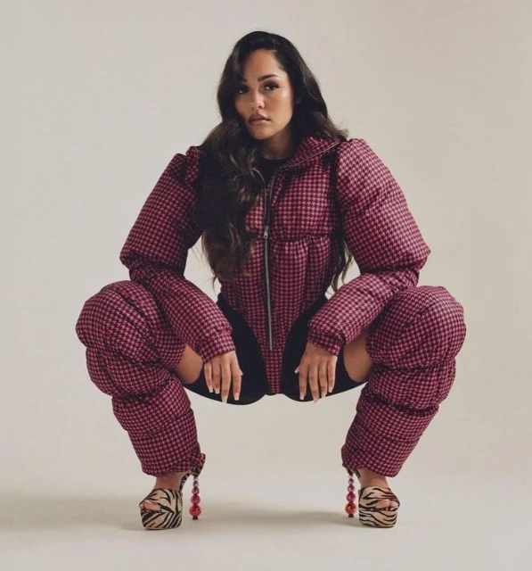 A woman squats in a studio wearing a puffy red and black houndstooth jacket with matching leg coverings and platform heels with beaded details.