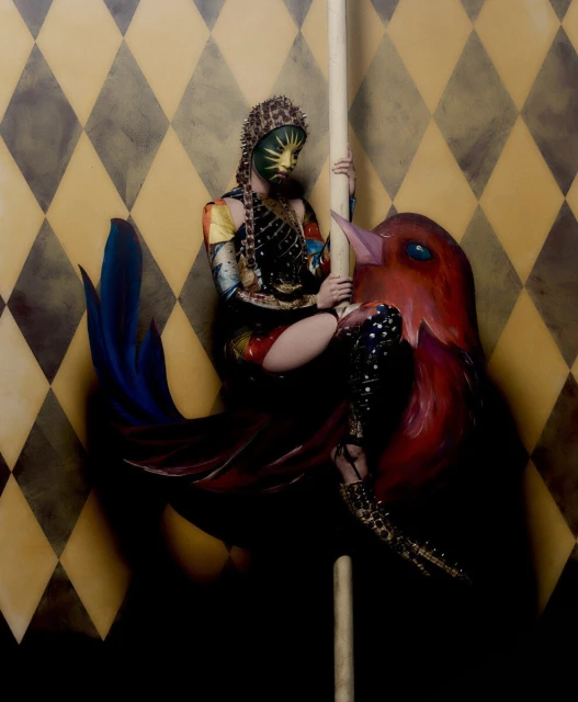 A woman in colorful, avant-garde attire sits atop a painted bird sculpture with a diamond-patterned backdrop, creating a surreal and artistic image.