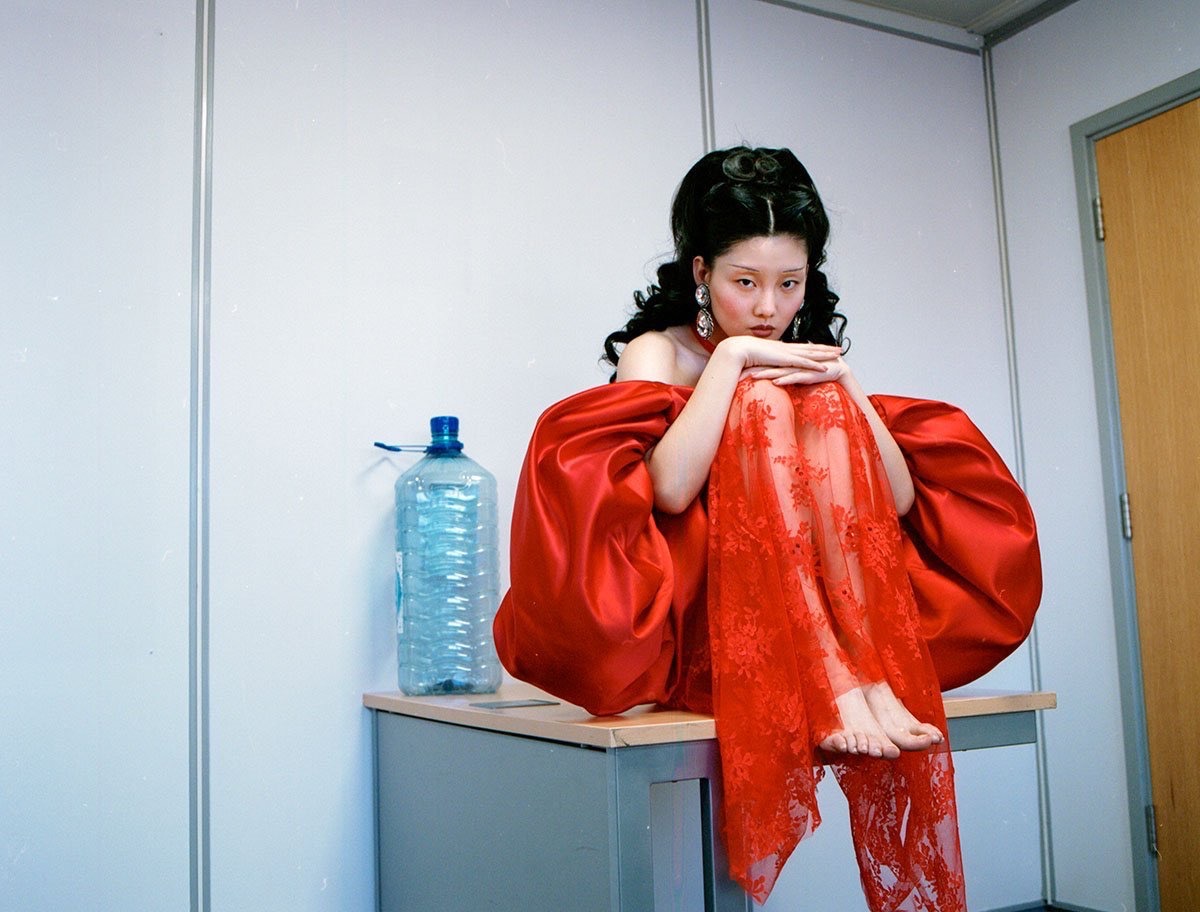 A young woman with dark hair in a red dress sits on a grey desk in front of a white wall with a door and a large water bottle.