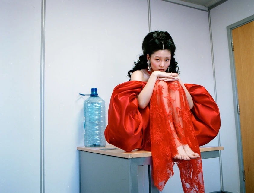 A young woman with dark hair in a red dress sits on a grey desk in front of a white wall with a door and a large water bottle.