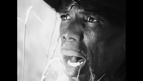 Close-up black and white portrait of a Black man with an open mouth, looking distressed, set against a blurred, light-toned background.