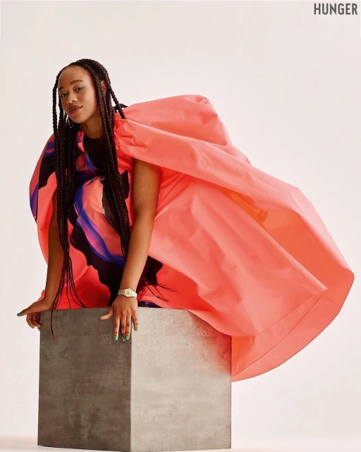 A young woman with long braids poses in a coral dress on a gray cube against a white backdrop in a studio fashion shot.