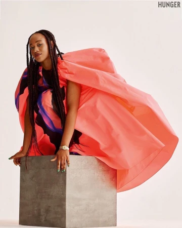 A young woman with long braids poses in a coral dress on a gray cube against a white backdrop in a studio fashion shot.