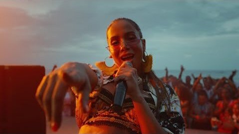 A woman sings into a microphone on the beach in front of a large crowd at sunset, illuminated by warm light and wearing large earrings.