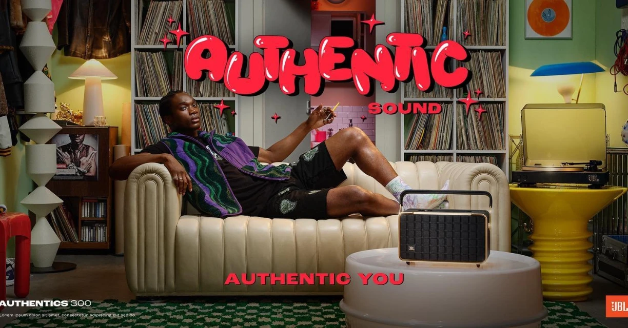 Black male model relaxes on a beige couch, listening to music on a JBL Authentics 300 speaker in a vibrant retro-styled room.