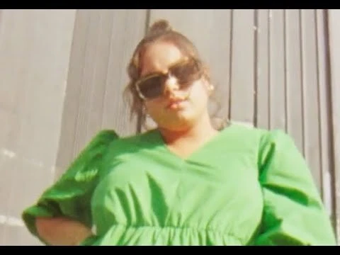 Portrait of a woman in a green dress and sunglasses, standing outdoors with a neutral expression, and a blurred background.