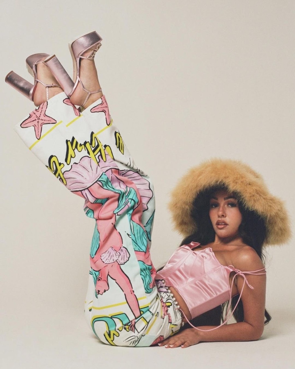 Woman in retro-inspired patterned pants and furry hat, posing indoors. Bright, soft lighting, high-heeled shoes, and light pink top.