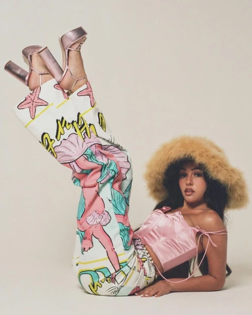 Woman in retro-inspired patterned pants and furry hat, posing indoors. Bright, soft lighting, high-heeled shoes, and light pink top.
