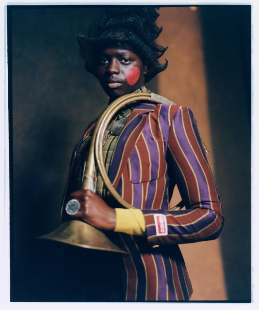 Portrait of a person with a painted heart, wearing a striped suit and hat, holding a gold-colored horn against a neutral backdrop.