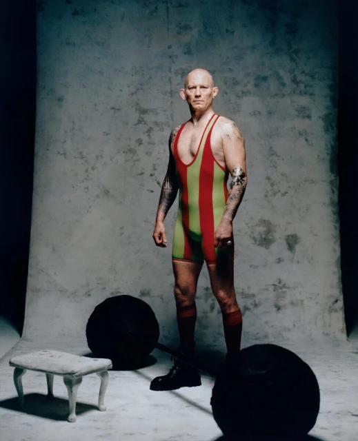 A bald adult male in a striped wrestling singlet stands next to a barbell weight and stool in a photo studio with a gray backdrop.