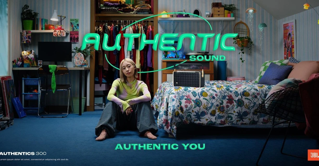 Adult woman poses next to portable speaker in a decorated bedroom with desk, closet, bed, blue carpet, and neon word art.