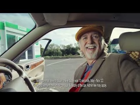 Smiling older man with a white beard and a yellow hat inside of a car at a gas station on a cloudy day, seen from the passenger seat.