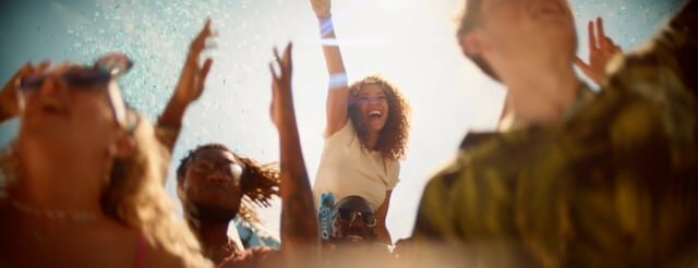 Diverse group of young adults enjoy a sunny outdoor gathering, raising their arms with joyful expressions; one woman is elevated on another's shoulders.
