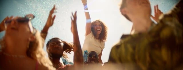Diverse group of young adults enjoy a sunny outdoor gathering, raising their arms with joyful expressions; one woman is elevated on another's shoulders.