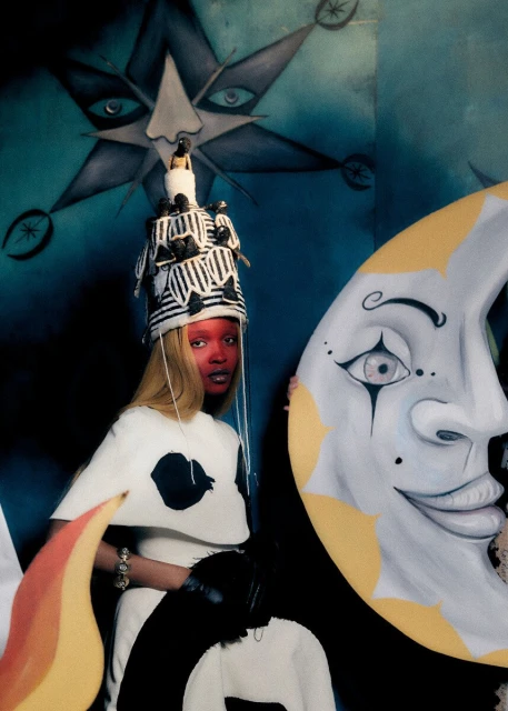 Artistic portrait of a person with painted face, wearing a patterned hat and white dress, posing next to a moon prop in a studio setting.