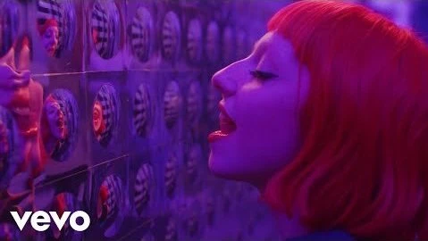 Profile shot of a woman with red hair standing in front of a wall covered in small, distorted mirror images, reflecting neon purple lighting.