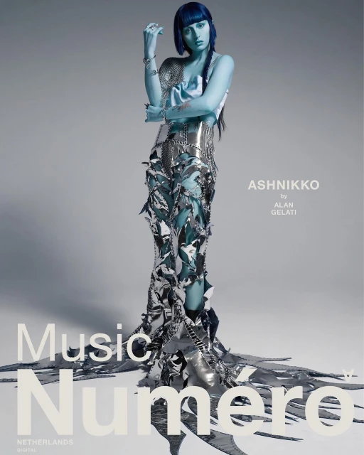 A full-length studio shot of a female model with blue skin and hair wearing an avant-garde silver dress with cut-out details.