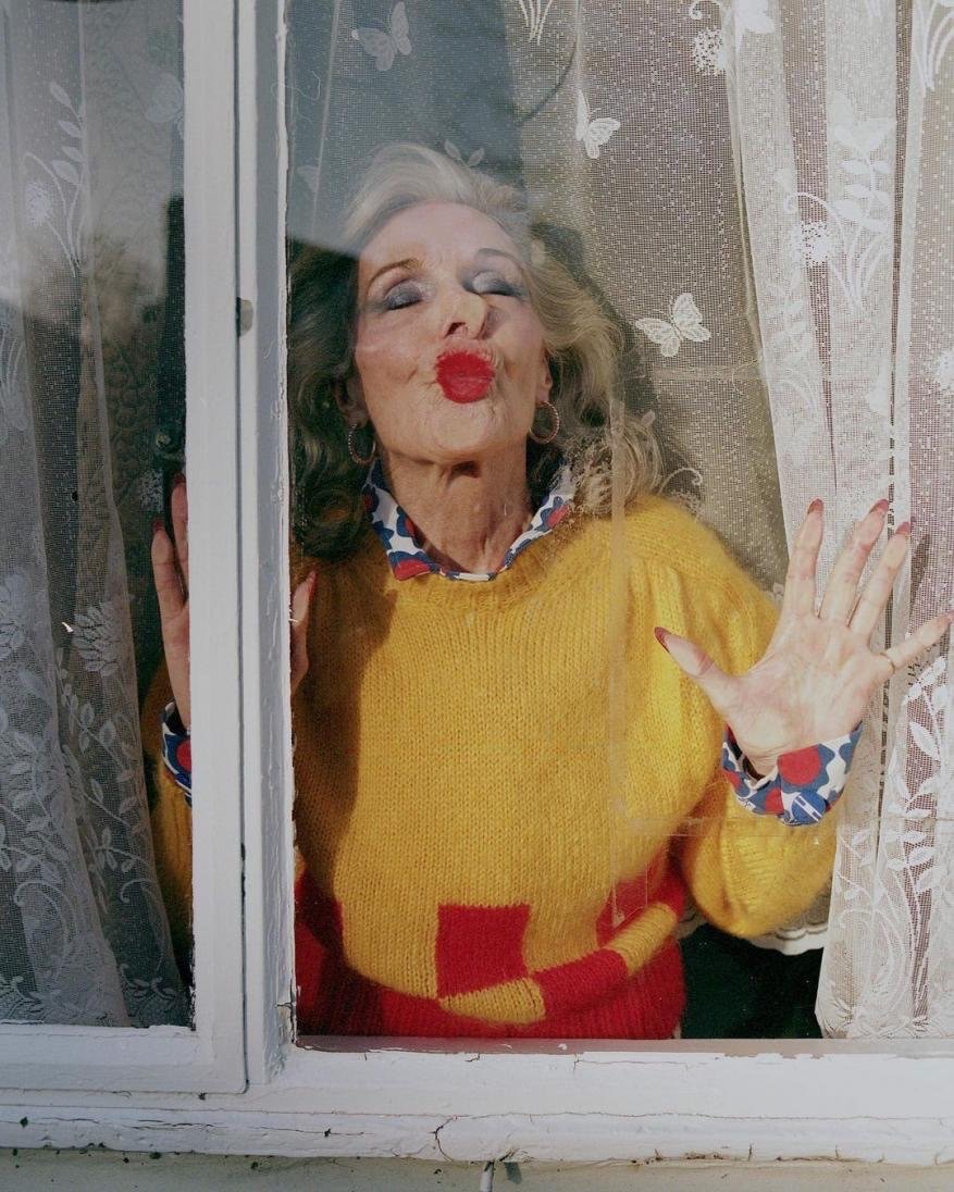 Elderly woman with red lipstick in yellow sweater, making a kissing face from behind a window with butterfly curtains. 
