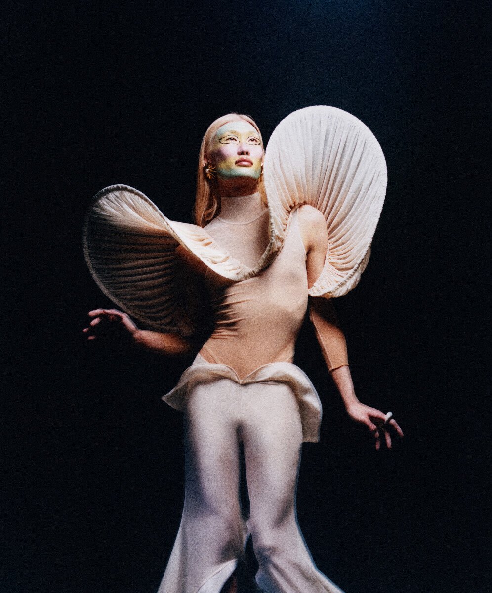 Person in Avant-Garde cream couture dress and neutral body suit with theatrical face makeup in a low angle fashion studio shot.