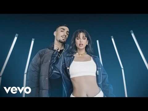 Music video still featuring a young adult couple: a woman in a white top and jeans jacket with a man in a puffer jacket, in a studio.