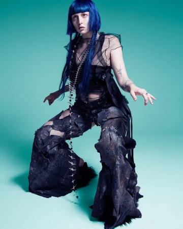 Model with vibrant blue hair wearing a sheer black top and wide dark pants against a teal background