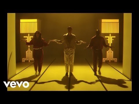 Three people dancing in a studio with yellow lighting and geometric patterns in the background, captured in a medium shot.