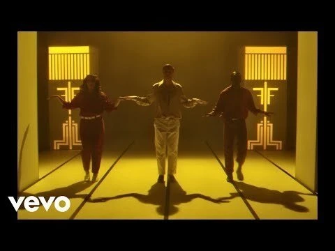 Three people dancing in a studio with yellow lighting and geometric patterns in the background, captured in a medium shot.