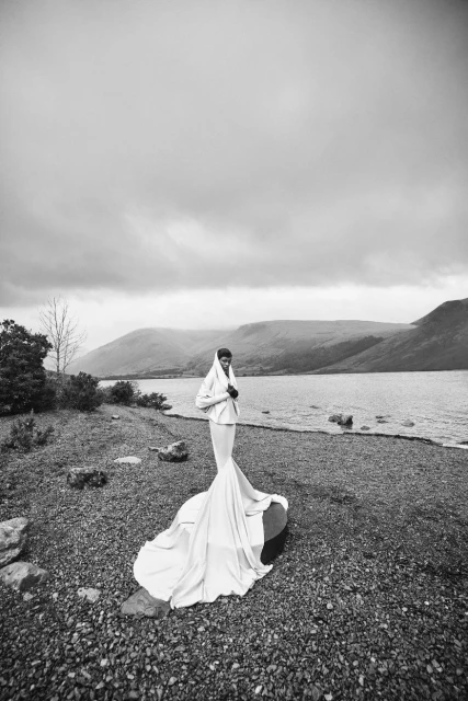 A woman in a white gown stands on a rocky shore near a lake and mountains under a cloudy sky in a black and white editorial fashion shot.