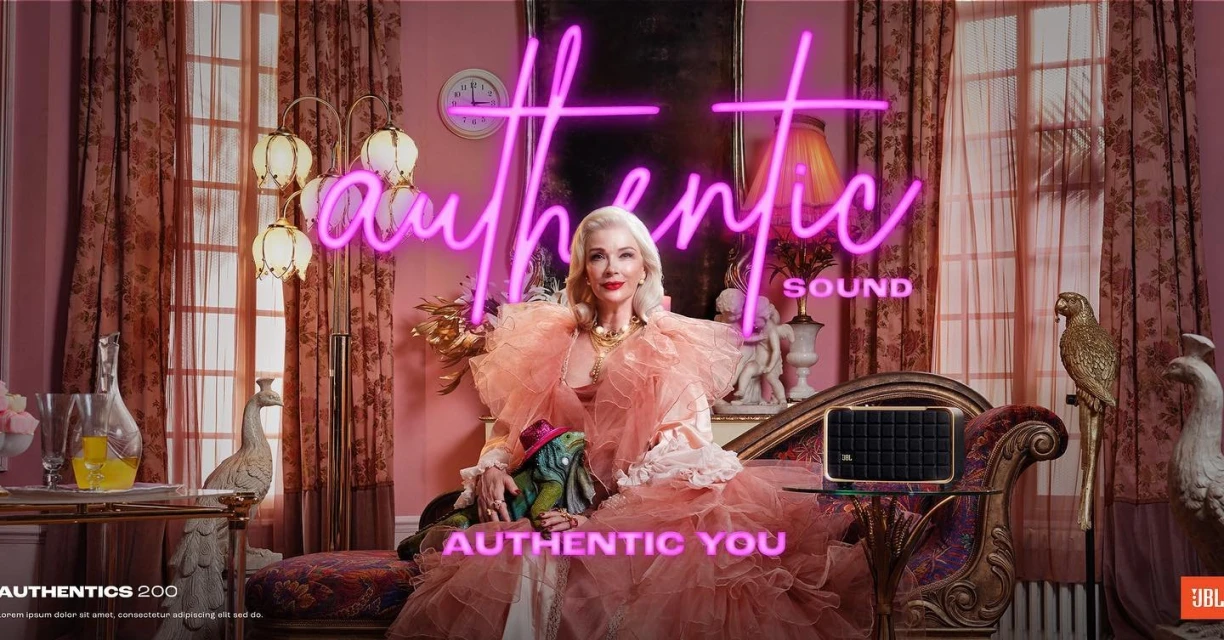 Elegant older woman in a pink dress sits with a lizard, promoting JBL Authentics speaker, illuminated by a neon sign in a pink interior.