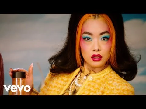 A woman with orange hair and colorful makeup wearing a yellow jacket poses in front of a cloudy background with a small container.