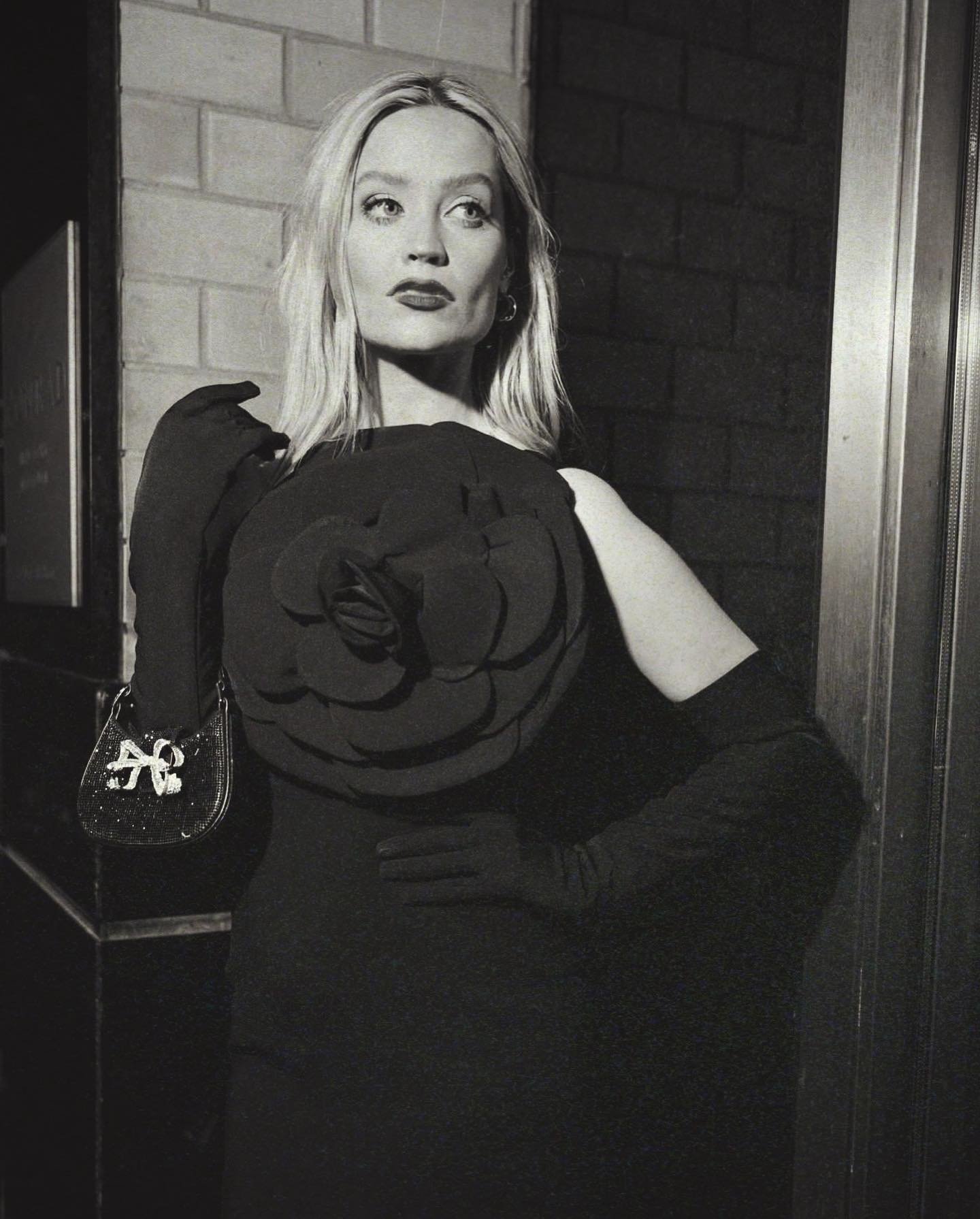 Black-and-white studio portrait of a blonde woman in a black dress with floral detail, carrying a purse and posing elegantly by a brick wall.