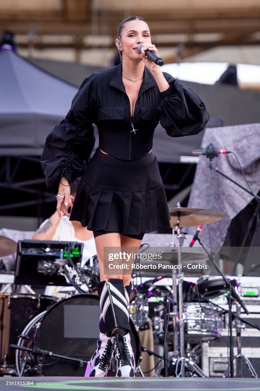 A singer performs on stage with a microphone, wearing a black outfit with inflated sleeves and knee-high boots, in front of musical equipment.