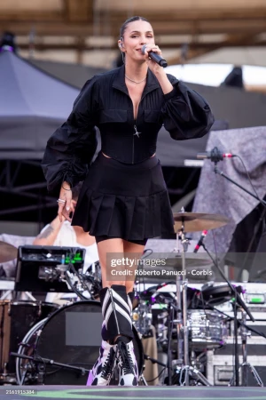 A singer performs on stage with a microphone, wearing a black outfit with inflated sleeves and knee-high boots, in front of musical equipment.