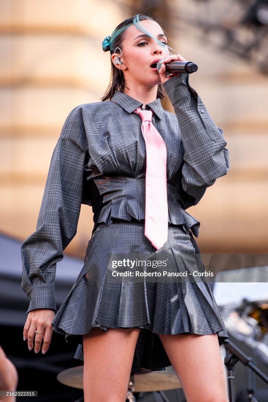 A singing female musician performs on outdoor stage wearing gray plaid outfit, pink tie, and turquoise hair accessories into a microphone.