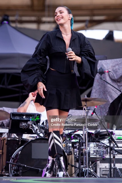 A woman with blue-tipped hair sings on stage wearing a black jacket and skirt, backed by a drummer and mics on an outdoor concert stage.