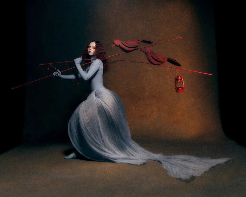 A pale woman in an extravagant blue gown strikes a pose, shouldering a wooden contraption with red birds and a red lantern in a studio setting.