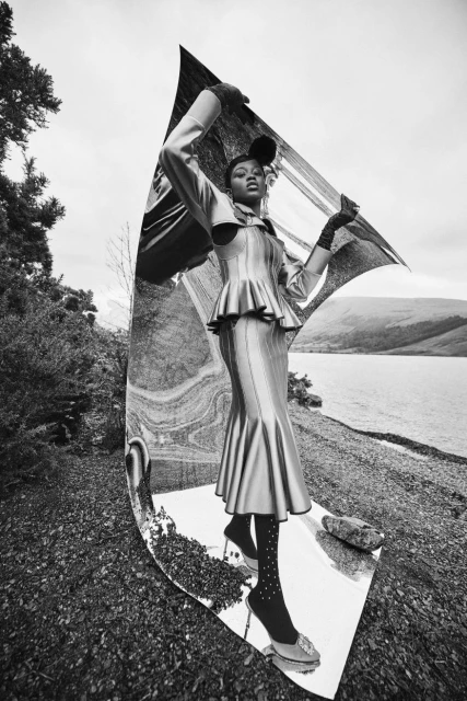 A person in a metallic dress poses with a mirror in a natural setting, wearing heels and polka dot socks. Mountains are in the background.