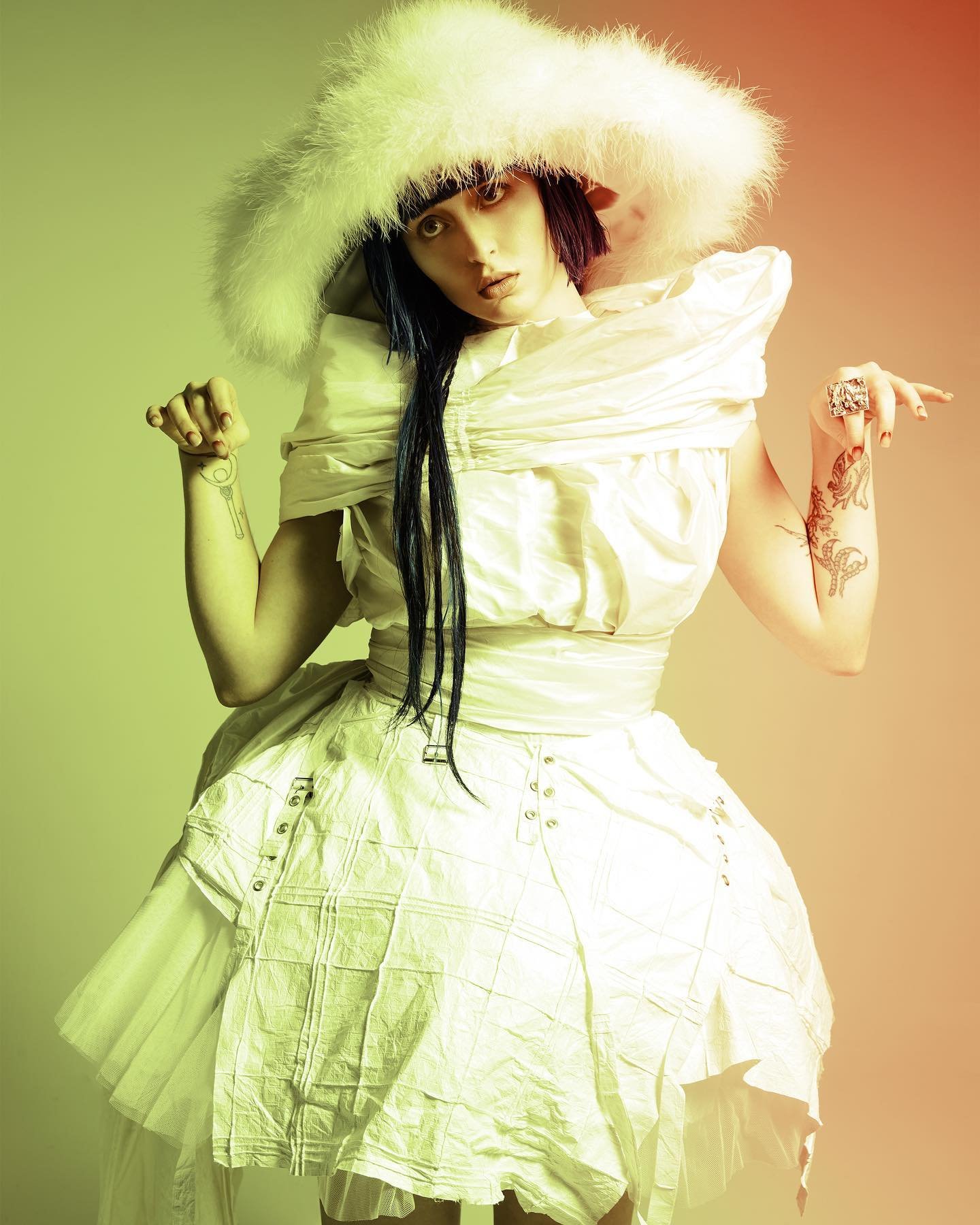 Woman in sculptural white dress and white fur hat strikes a pose in a studio with a soft, diffused lighting and a warm color gradient.