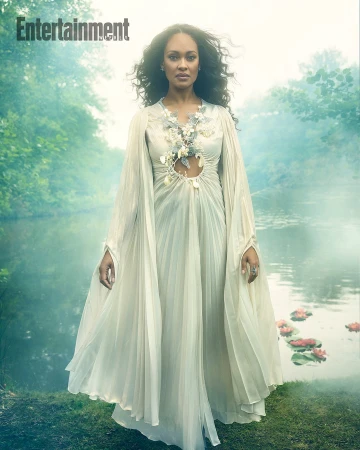 Adult female with curly hair poses in a white dress with floral details in a hazy outdoor setting near water and greenery.