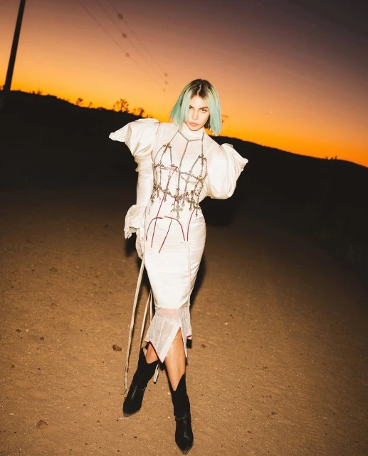 Woman in a white couture dress and black cowboy boots stands in a desert landscape at sunset, creating a high-fashion editorial style image.