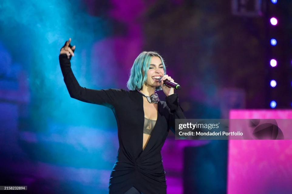 A singer with teal hair performs on stage, holding a microphone and wearing a black dress with a statement necklace under bright spotlights.